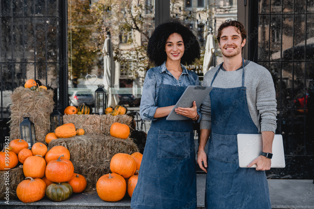 Wall mural happy young coffee shop owners standing outdoor near their cafe with laptop and decorating pumpkins 