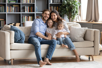 Portrait of overjoyed young Caucasian family with two little daughters sit rest on cozy couch in living room. Happy parents and small girls children rest on comfortable sofa enjoy weekend at home.