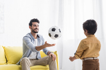 Smiling hispanic man throwing football to son at home, two generations of men