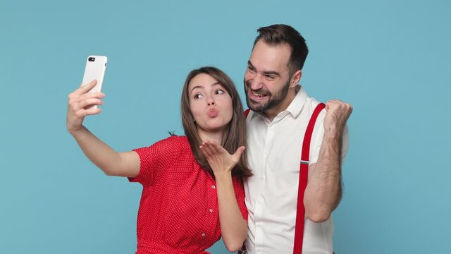 Funny young couple friends man woman in white red clothes posing isolated on blue background. People lifestyle concept. Doing selfie shot on mobile phone showing victory sign thumb up winner gesture