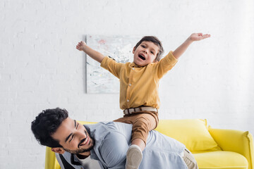 Cheerful boy sitting on back of hispanic father in living room, two generations of men