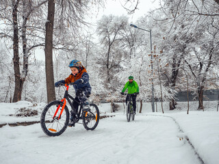 A boy and his father ride bicycles in a winter park. Wonderful winter day outdoors after snowfall. The bearded man is the boy's father, follows him on a bicycle.