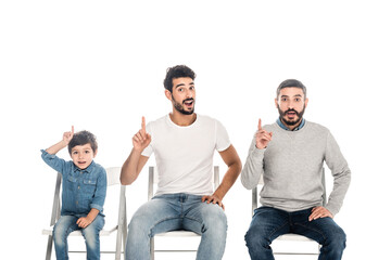 amazed boy with hispanic father and grandpa pointing with fingers while sitting on chairs isolated on white, three generations of men