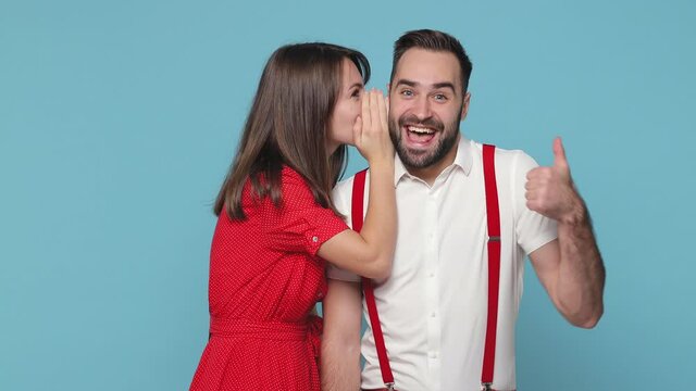 Excited young couple friends man woman 20s in white red clothes isolated on blue background studio. People lifestyle concept. Whispering gossip tells secret behind hand showing thumb up winner gesture