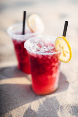 Tropical red ice cold drinks. A glass of cold drink stands on the sand. Drinking straw and orange slice. Hot summer and relaxation.
