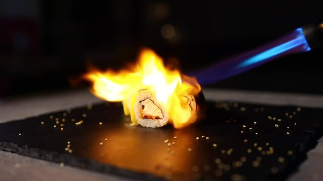 The process of baking Japanese sushi lying on a black iron sheet surrounded by sesame seeds with a gas burner. Close-up