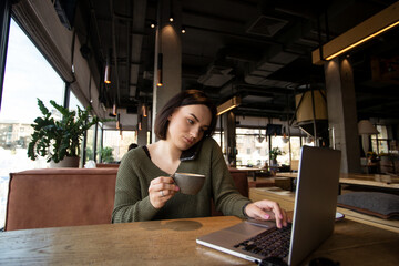 Pretty young woman works at laptop in a cozy coffee shop with big bright windows on background. Female freelancer presses smartphone to shoulder with her head to speak and holds cup of coffee.