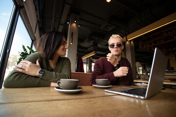 Couple working together in cafe. Young brunette woman smiling and looking at cool blonde man in black round glasses. Gray laptop on table. Two cups of coffee. Freelance working concept.