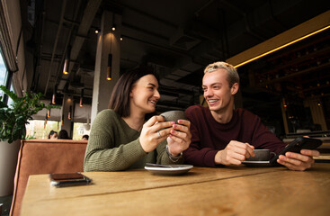 Caucasian couple drinks coffee in cafe. Brunette woman warms her hands on the cup she holds in her hands and smiles to blonde man in purple sweatshirt. Cozy atmosphere in coffee shop.