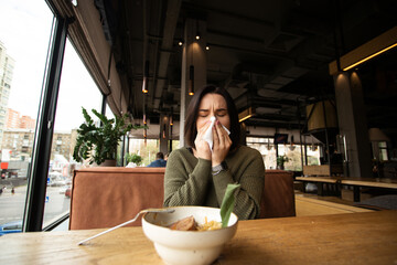 Sick young woman with runny nose came to a cafe. Public place on bakground. Brunette woman blows her nose. Isolate yourself if you get sick. Restaurant or cafe concept.