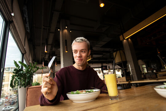 Very tasty caesar calad Cheerful young man eating healthy food in restaurant and smiling. Glass of orange fresh. Public place on background. Stylish young man tasting delicious meal.