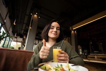 Tasty meal in the restaurant. Cheerful brunette woman shows thumb up gesture with one hand and holds a glass of orange juice in another. Bowl of caesar salad on foreground.