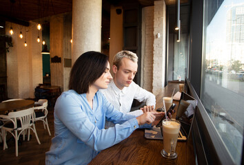 Two freelance workers in coffee house. Brunette woman showing something in her smartphone and pointing on screen, man listening. Decision making concept. Cup of latte and gray laptop on table.