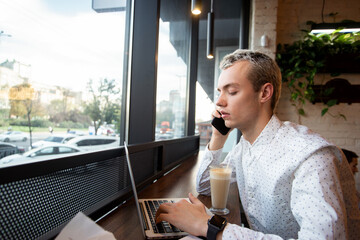 Man prints on modern laptop. Crispy croissant and cup of yummy latte. Cafe or restaurant on background. Young man talking on the phone in a public place.