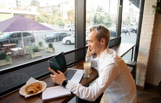 Young freelancer happy with a phone call. Remote work concept. Coffee house on background. Tasty croissant and cup of latte. Male freelancer programmer working in a coffee shop.