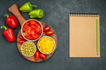 top view of bowls of vegetables on plate stand with vegetables and notepadon side on dark grey background