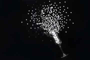 Fototapeta premium Silver star confetti and party streamers poured out champagne glass in flat lay style on black background. Christmas and New Year holiday celebration concept.