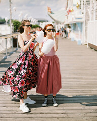Happy girls with sunglasses and prom dresses eating cotton candy at amusement park. Teenagers in pink evening dresses on vacation. Birthday party idea. Summer holiday.