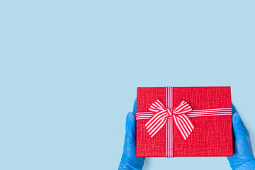 Fototapeta na wymiar Hands in blue medical gloves hold a red gift box with ribbon isolated on blue background, copy space. Can be used to illustrate safe gifts, protect loved ones, gifts during the coronavirus pandemic