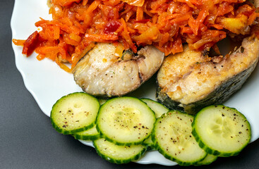 Top view of fried cod fish slices, fresh cucumbers and stewed onion with carrots on the white dinner plate.