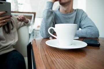White cup of coffe on front. Blurred couple on background. Man and woman on romantic date in cafe. Coffeehouse. Woman holding her smartphone and texting something when man waiting for her to finish.