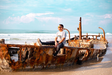 Young man sits on the hull of an old destroyed ship on the seashore. Looks info the distance,...