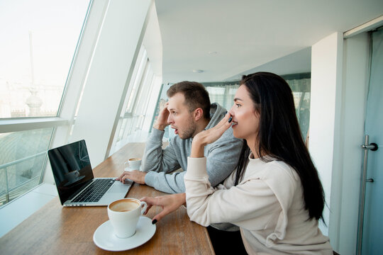 Couple saw something shoking on laptop. Man fiddles with hair and woman giggles and covers her mouth with hand. Empty coffee cup. White light room on background.