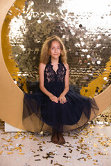 A cute teenage girl in a beautiful dress sits in a New Year's photo zone in the form of a golden moon