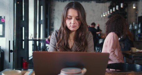 Portrait of young Caucasian brunette female freelancer using laptop for studies and work at busy loft coworking cafe.