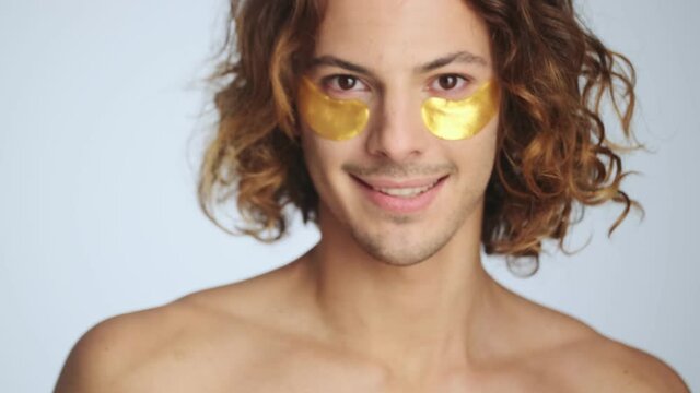 A happy man is wearing hydrogel eye-patches standing isolated over white wall in studio