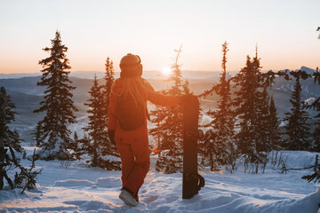Female with snowboard. Snowboarder standing and resting in winter forest watching beautiful sunset...
