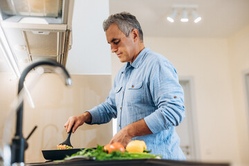 Mature man in blue shirt cooking on modern kitchen. Light cozy room on background. Concentrated man standing by stove, stews vegetables in a frying pan. Healthy food. Bright vegetables on front.