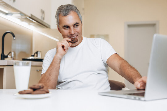 Mature Man Sitting On Kitchen, Using His Modern Gray Laptop, Watching Funny Series Or Videos, Laughing And Eating Yummy Cookies. Glass Of Fresh Milk And Biscuits On White Table.