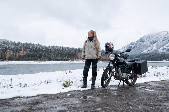 Female motorcyclist standing with her motorcycle near snowy mountain river, snow peaks skyline view