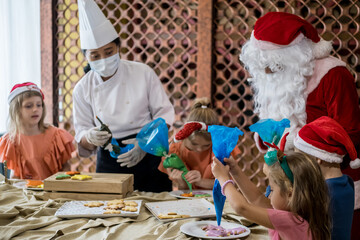 Children decorating Christmas biscuits and gingerbread cookies in kitchen together with chef and Santa Claus. Kids cooking class. Merry Christmas and happy holidays