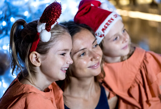 Single Mother And Two Daughters By The Christmas Tree Wearing Festive Hats, Laughing And Looking Away. Merry Christmas And Happy Holidays