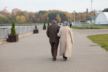 An elderly married couple of retirees walk together in an autumn park. View from the back.