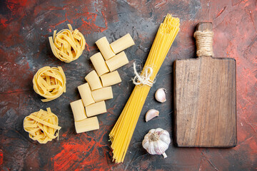 Top view of different types of uncooked pastas and wooden cutting board garlic on black background