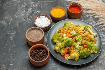 Side view of healthy meal with brocoli and carrots on a black plate and spices on gray background