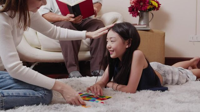 Asian mother teaching her pretty daughter to play games for developing skills and Asian retirement grandfather reading a book on sofa in the living room. Happy Family educational at home concept.