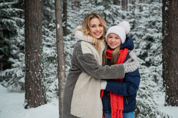 Fototapeta na wymiar Charming young blonde hugs her teenage daughter in winter in the woods, looking at the camera, portrait.