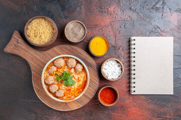 Above view of tomato meatballs soup with noodles in a brown bowl different spices and notebook on dark background