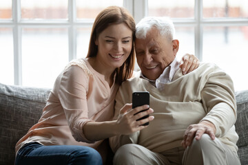 Look, grandpa. Loving adult granddaughter hugging smiling senior grandfather taking selfie with him on phone. Young woman grown daughter embracing older man father showing photos of grandchild on cell