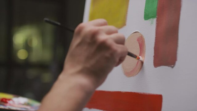 Close-up of unrecognizable male artist hand with paintbrush painting circle out of mixed white and red oil paints on canvas in dark empty art studio