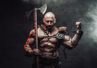 Wielding an axe and dressed in light armour with fur bald and furious viking warrior poses in dark and smokey background showing his huge biceps.