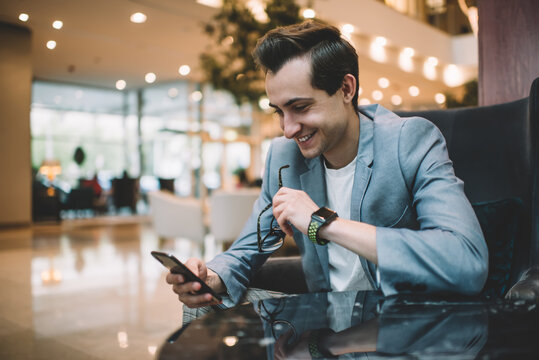 Cheerful successful male entrepreneur in formal wear satisfied with finance news checking mail on mobile phone, happy young 20s businessman satisfied with money transaction via smartphone application
