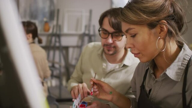 Slow-motion medium shot of young mixed-race woman at arts lesson learning basics of abstract painting listening to male teacher while loading paintbrush with gouache paint at bright studio