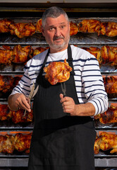 A seller in an apron offers grilled poultry at a weekly market. He has speared a rotisserie chicken...