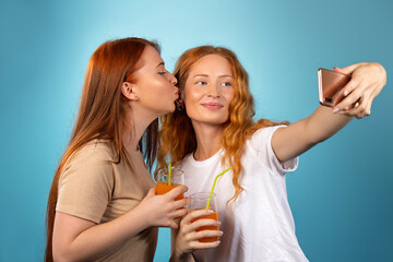 Redhaired female friends have a lovely time, drink juice and take selfies on a blue background. Online blogging