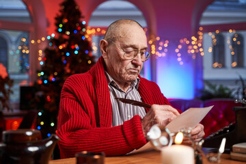 Portrait of a santa with glasses sitting at table and writing christmas cards with serious face